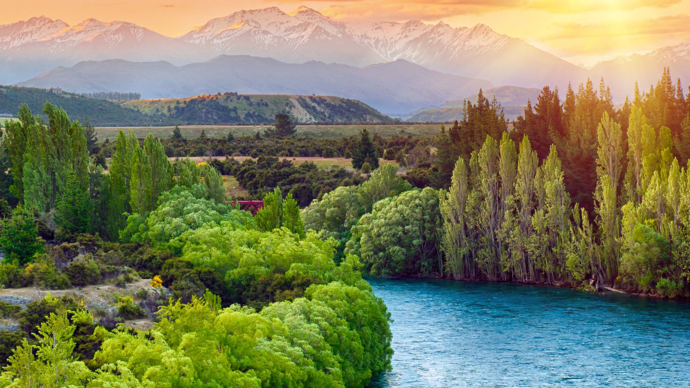 A river flows through a lush valley with mountains and a sunset behind