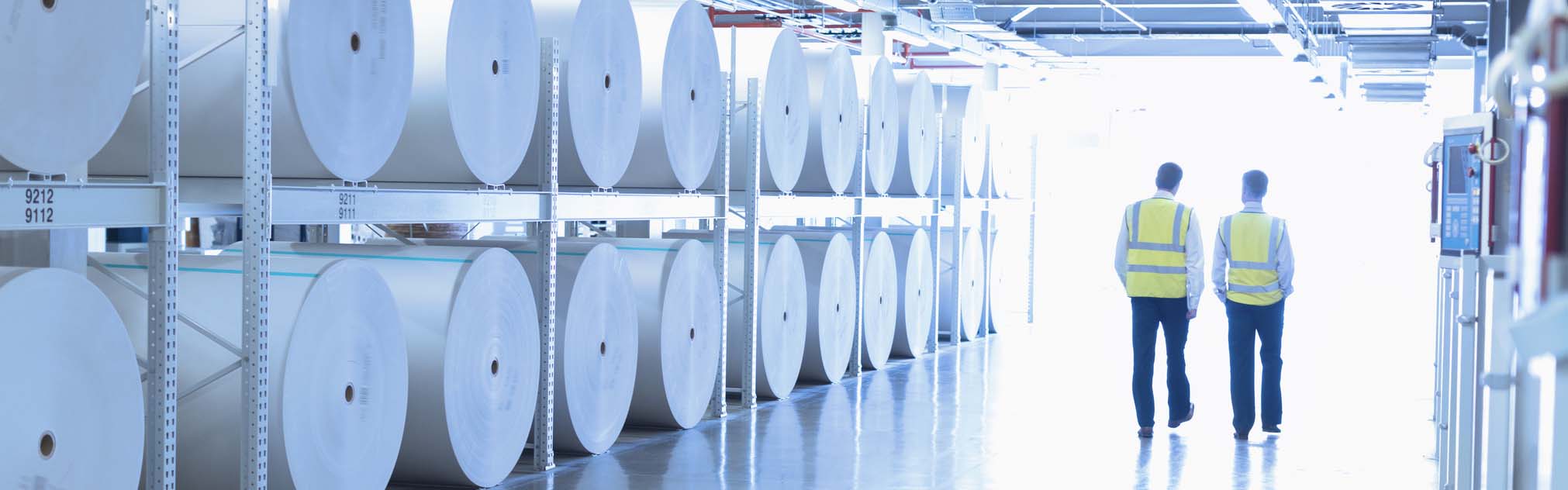 Pulp and paper mill workers walk down a hall next to reams of paper.